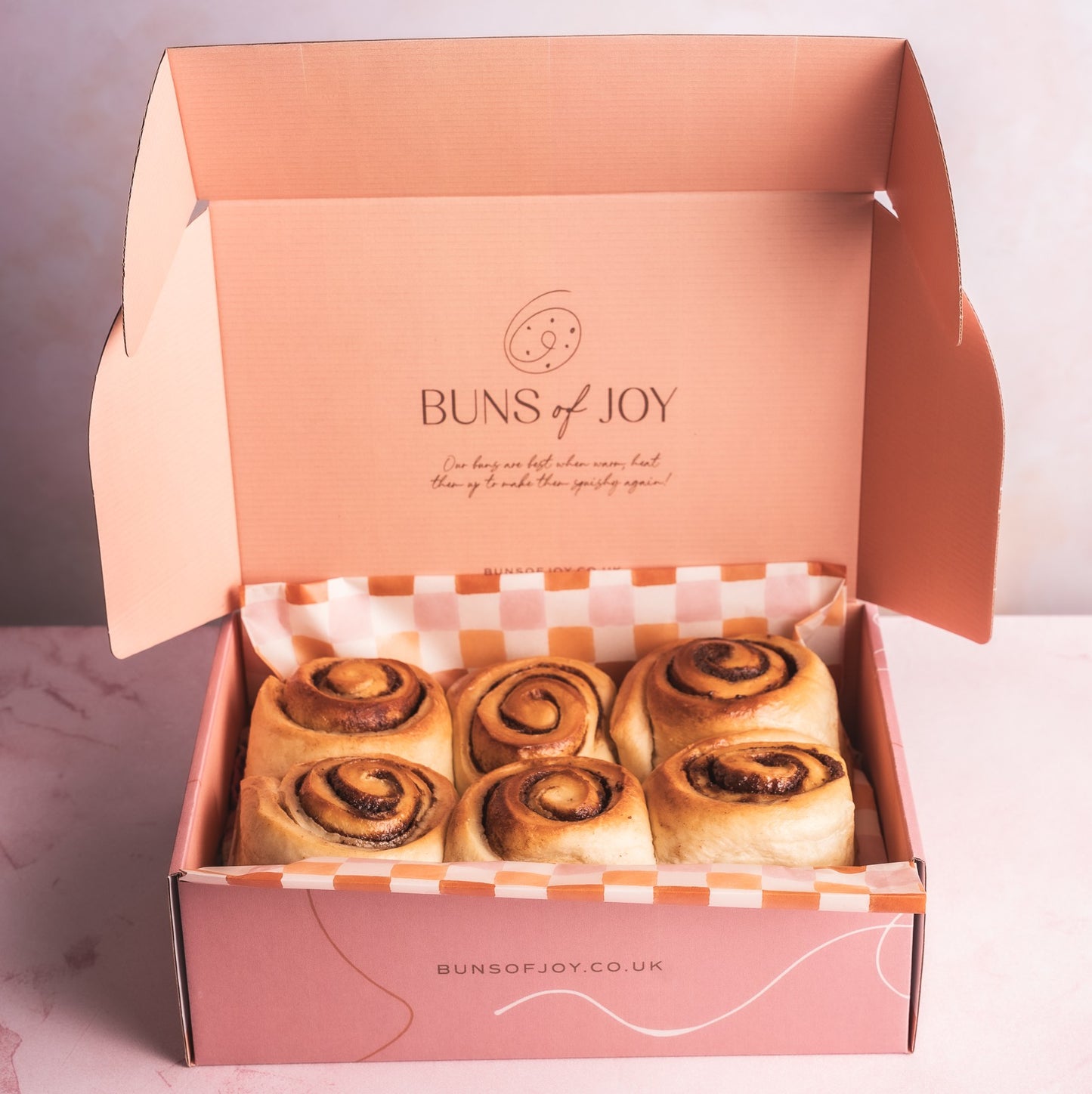 An open pink box of Glazed 'Buns of Joy' cinnamon buns on a light background