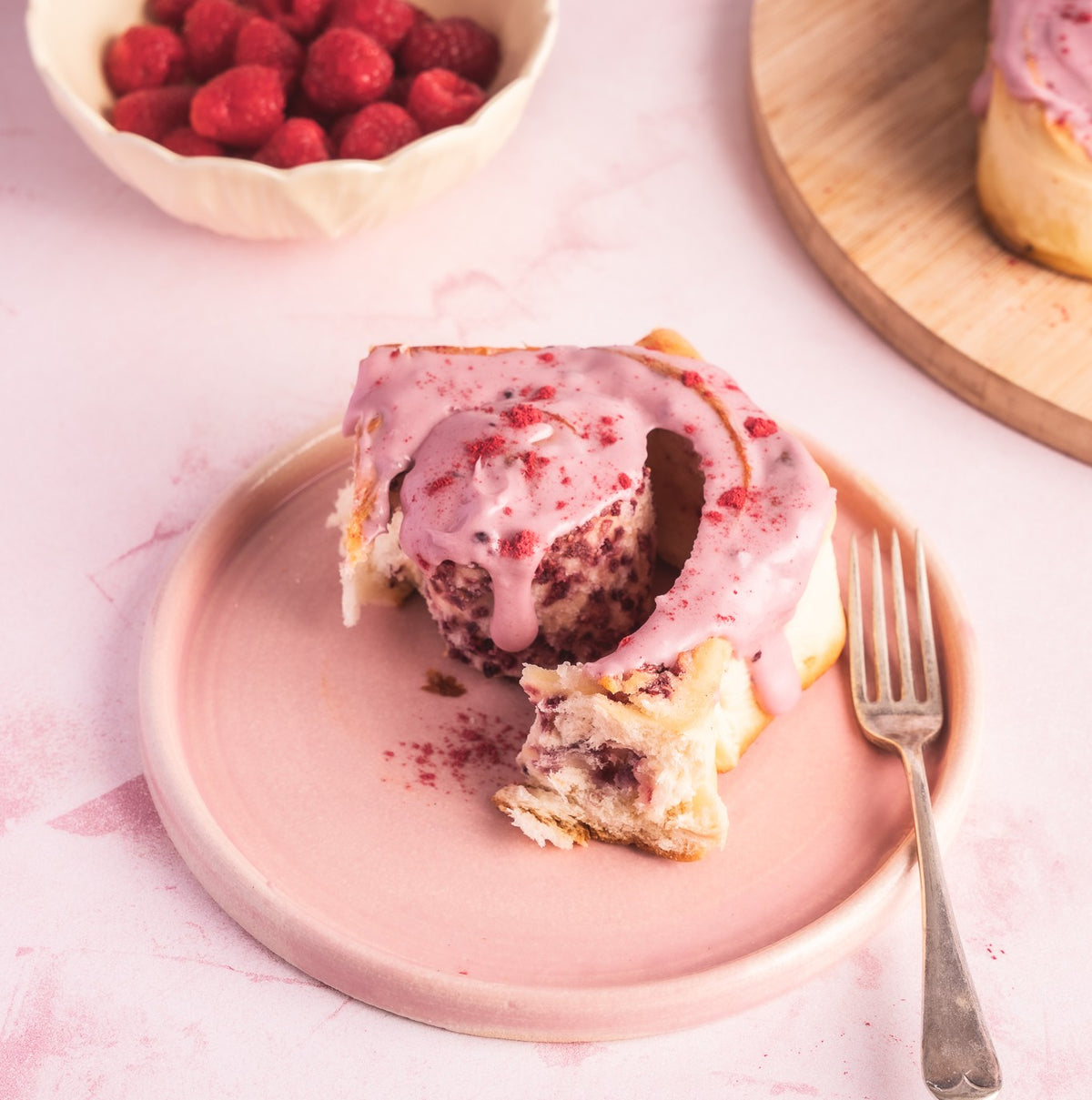 A Cinnamon Bun topped with pink frosting sits on a pink plate with a fork alongside and raspberries sit in a bowl at the back.