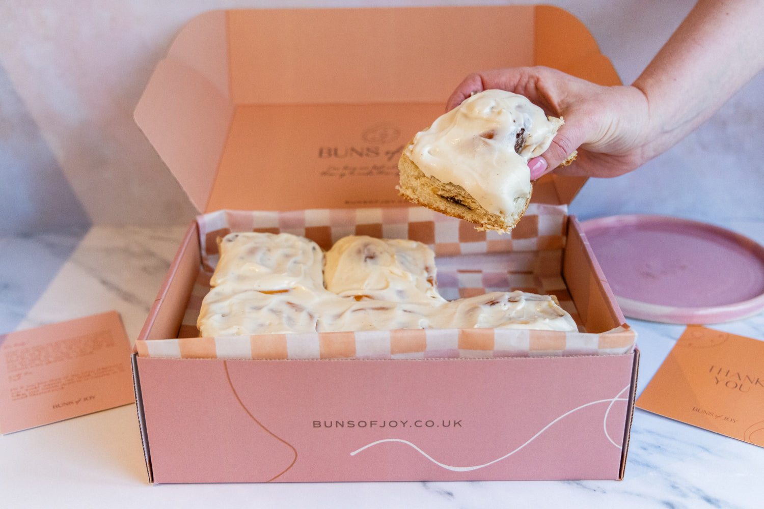 a pink box containing 6 delicious cinnamon buns topped with vanilla frosting