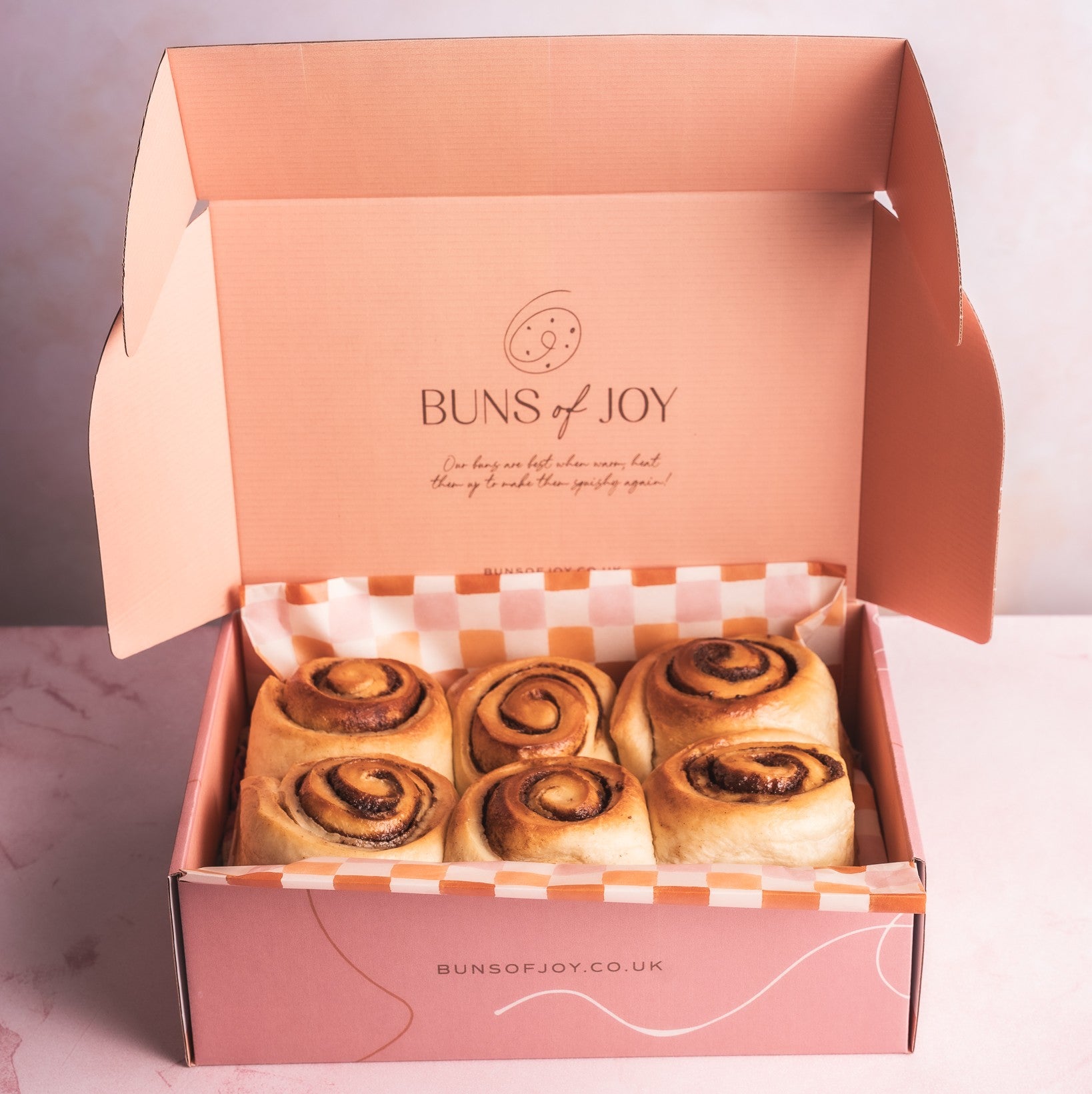 An open pink box of Glazed 'Buns of Joy' cinnamon buns on a light background