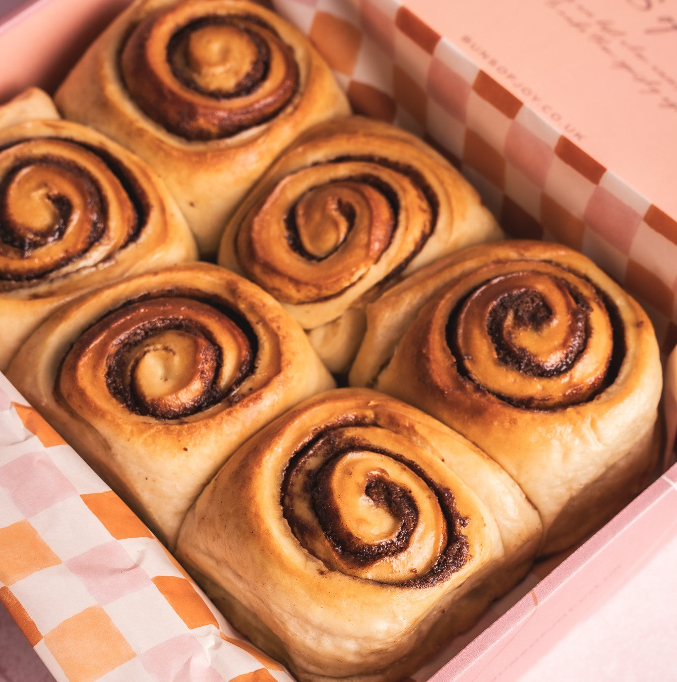 6 Glazed Cinnamon buns in a pink box with a checkered lining