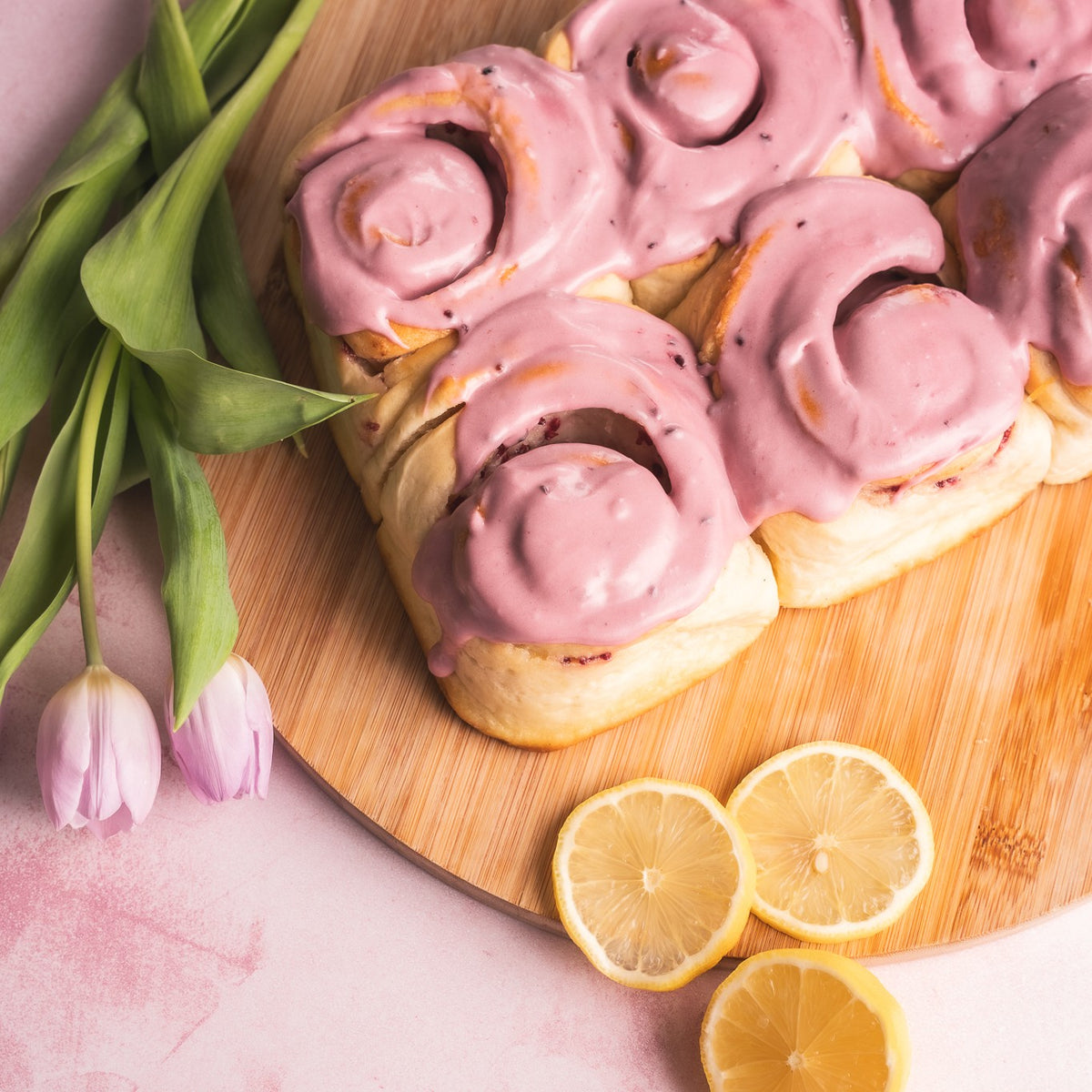 6 Cinnamon Buns topped with pink icing sit on a wooden board with tulips and lemons laid alongside.