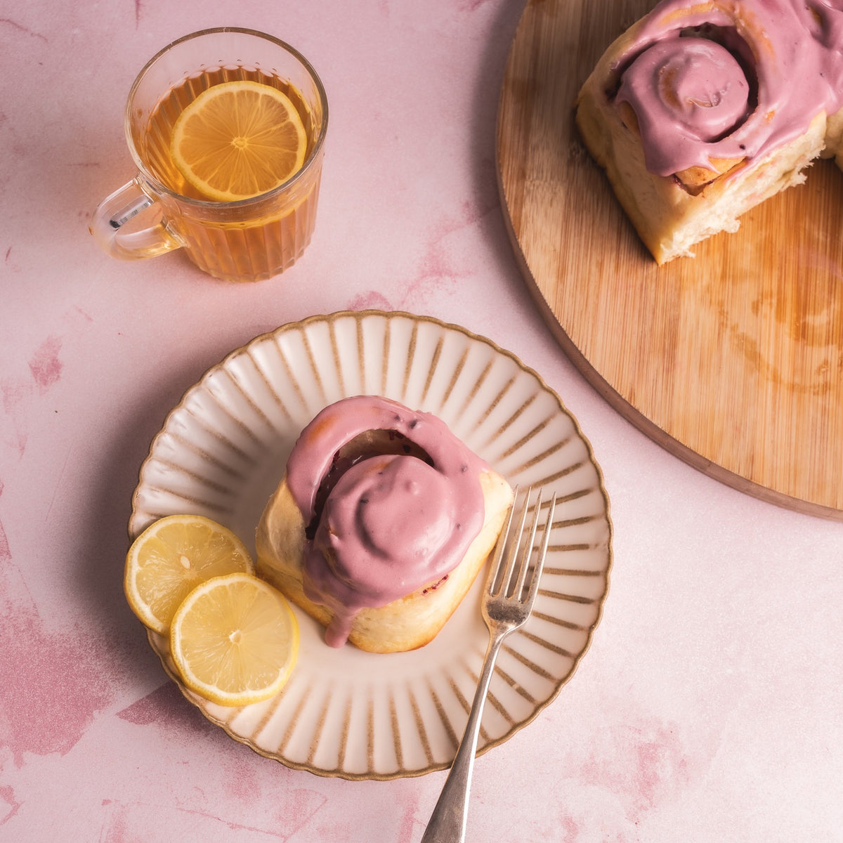 A Cinnamon Bun topped with pink frosting sits on a scalloped cream plate with lemon slices, next to a glass of iced tea and another cinnamon roll on a wooden board, all on a pink surface.